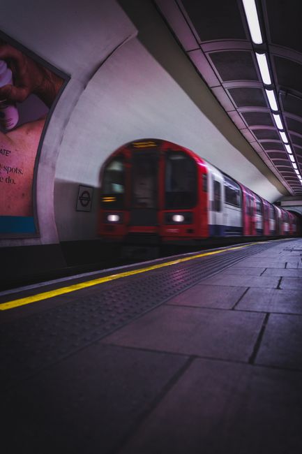 A blurred image of a train moving rapidly through an underground station platform, capturing the motion effect on the beige and dark brown exterior of the train. The platform surface features light-co
