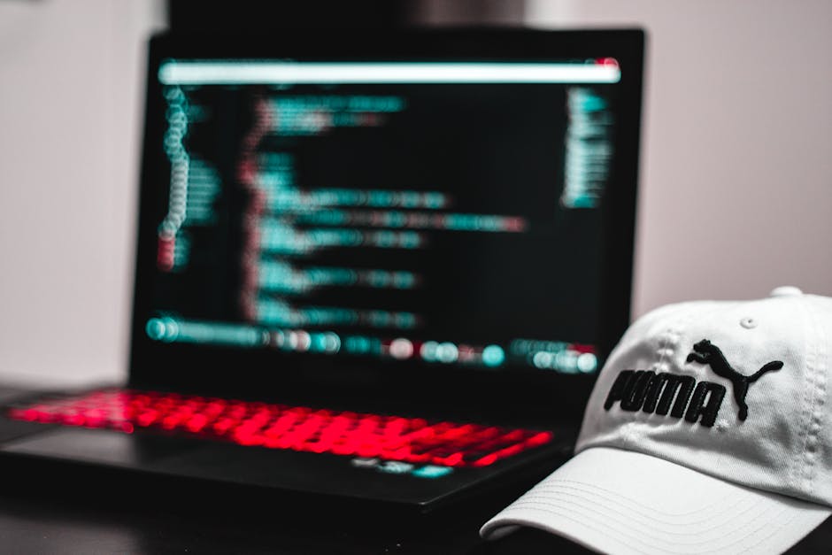 A close-up of a person's hands typing on a MacBook laptop positioned on a dark, reflective surface. The laptop screen displays lines of code in a text editor with a dark theme, featuring syntax highli